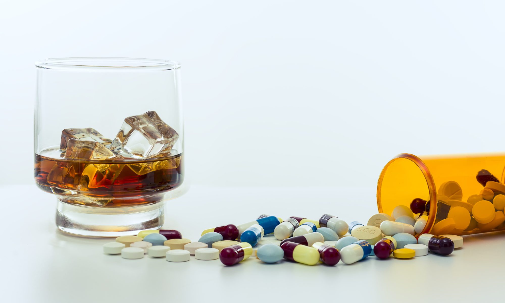 A glass of whiskey with ice next to a spilled prescription bottle and assorted pills on a white surface.