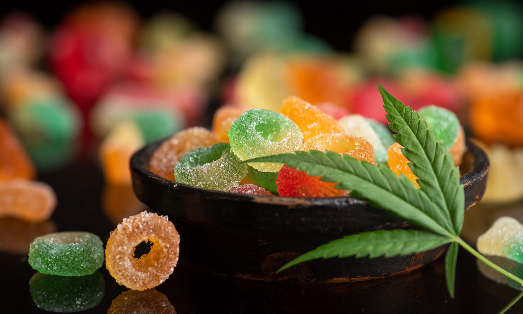 Assorted colorful cannabis gummies in a bowl with a marijuana leaf placed on top, symbolizing THC-infused edibles.