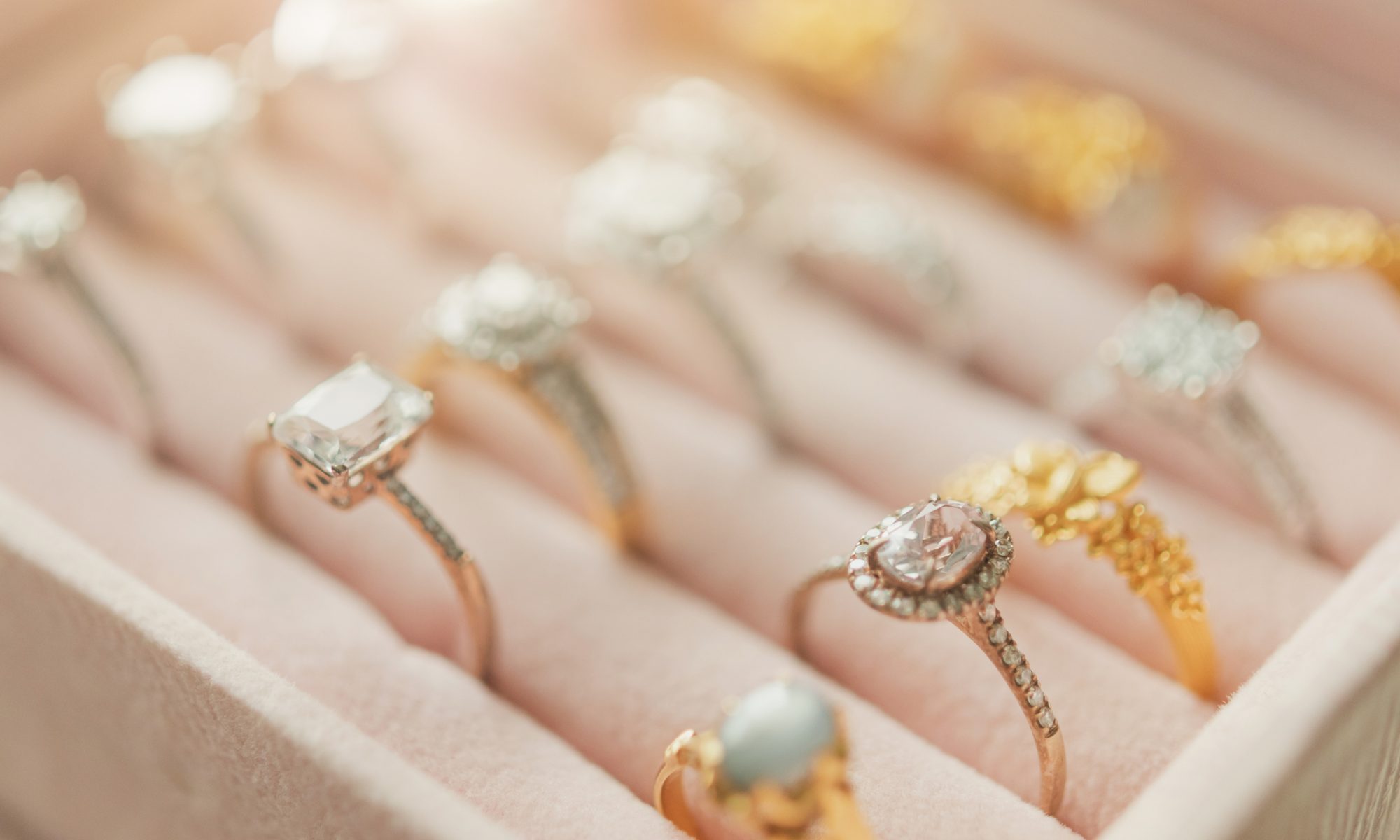 A display case of assorted gold and silver diamond rings arranged in neat rows on a soft pink velvet tray.