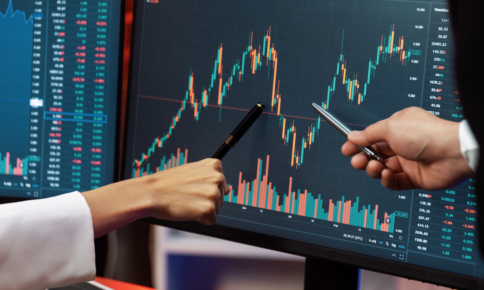 Close-up of two people pointing pens at a financial trading chart on a monitor showing rising and falling stock prices with colorful candlesticks and market data.
