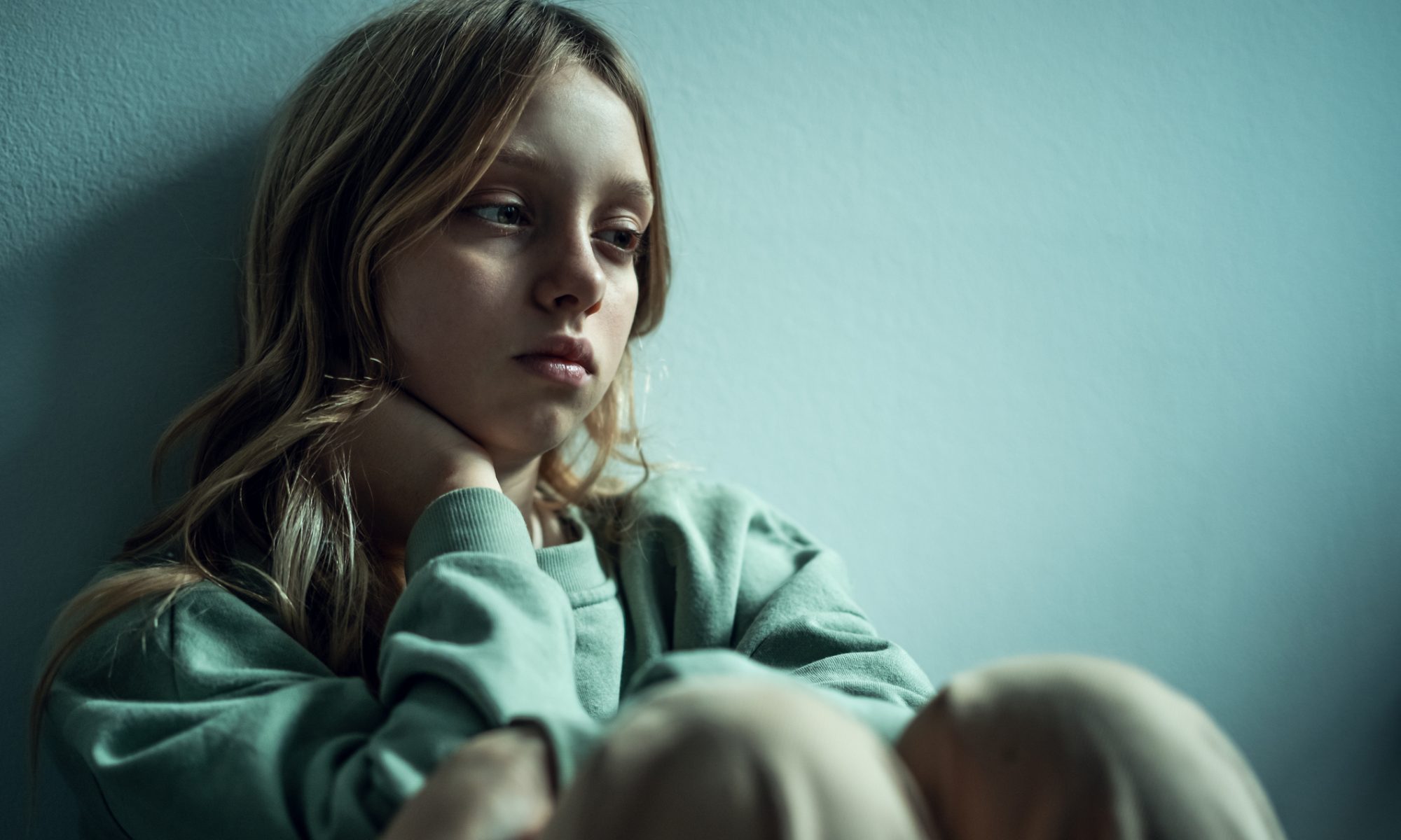 A sad adolescent girl sits alone against a wall, looking down with a troubled expression, symbolizing youth depression and mental health challenges.