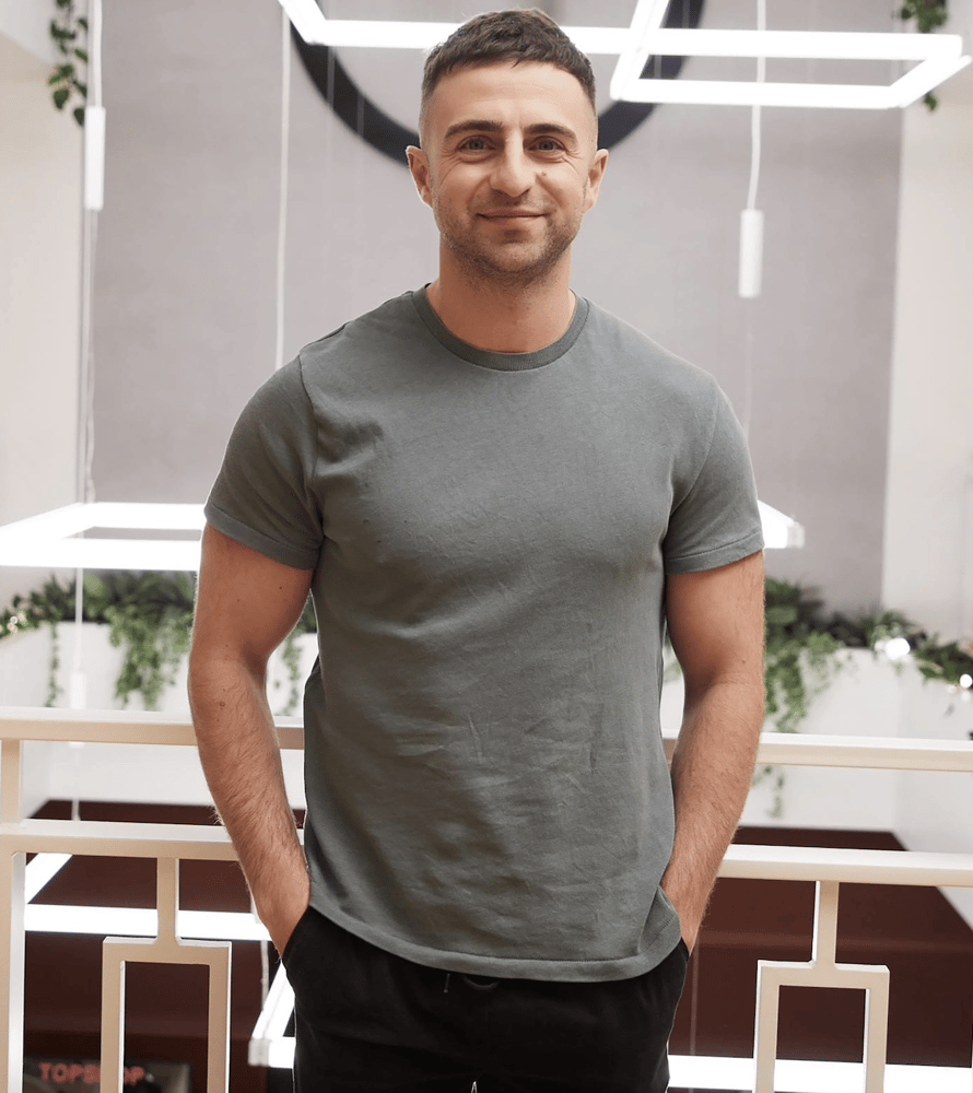 Sean Trend CFO of ALLSaints standing indoors, smiling, with his hands in his pockets. He’s wearing a fitted grey-green T-shirt and dark pants, and the background has modern geometric lighting and some greenery.