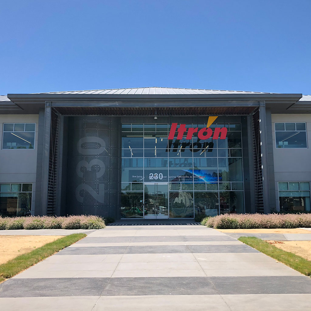 The exterior of a modern office building with large glass windows and the red and yellow “Itron” logo displayed above the main entrance. The building number “230” is visible on a vertical metal panel to the left of the doors. A wide walkway leads up to the entrance, bordered by neatly trimmed landscaping under a clear blue sky.