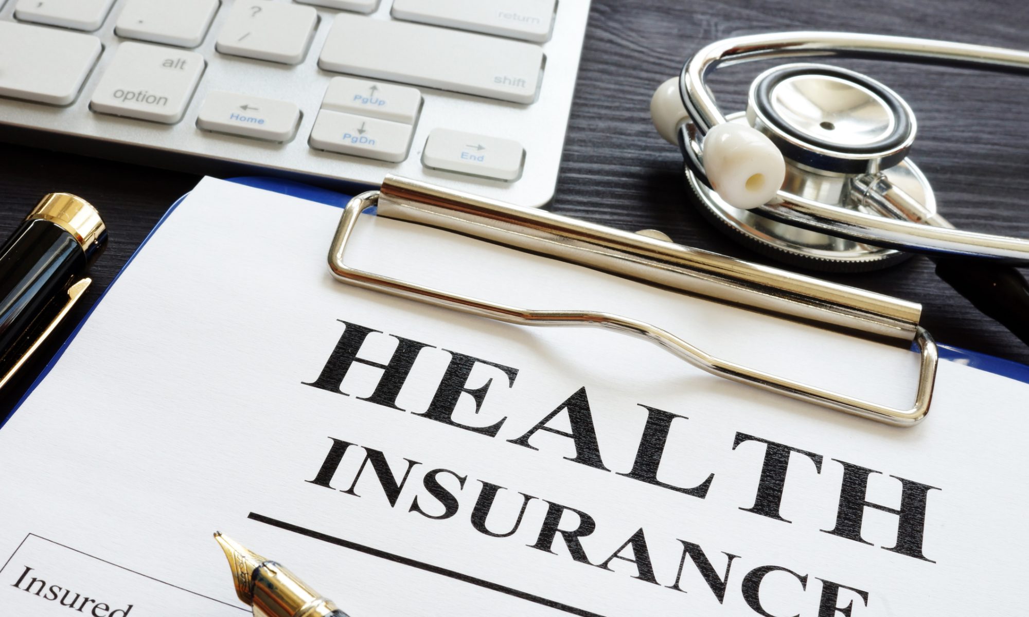 "A health insurance form on a clipboard placed on a desk, with a fountain pen, stethoscope, and part of a computer keyboard nearby."