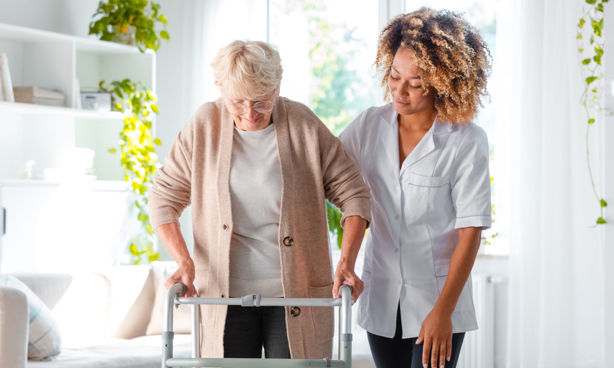 Elderly woman using a walker for support while a healthcare professional assists her in a bright, home-like care setting.