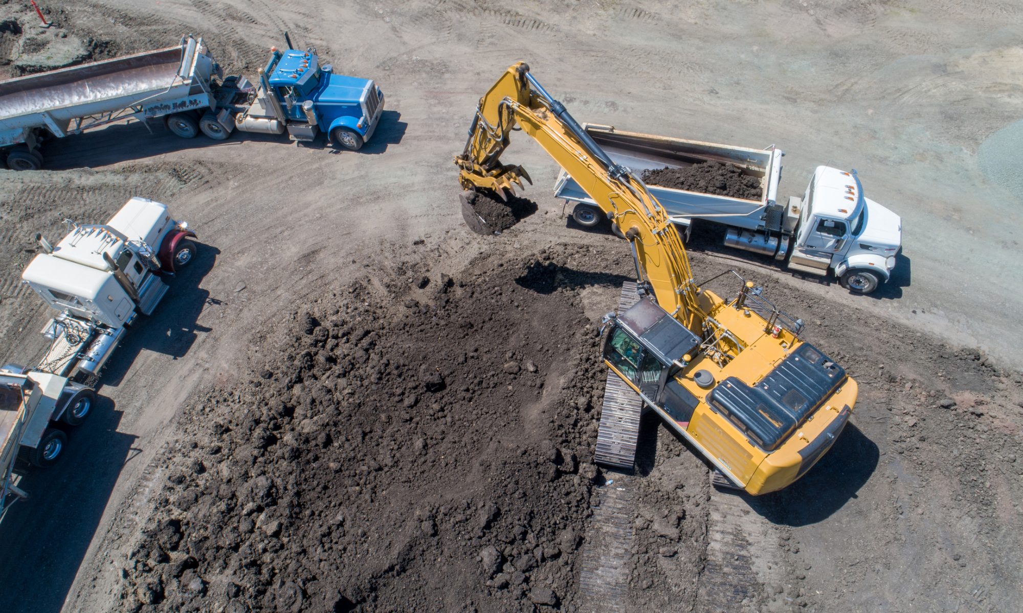 Aerial view of a construction site with heavy machinery, including an excavator loading soil into large dump trucks, as crews work on developing an industrial processing facility.