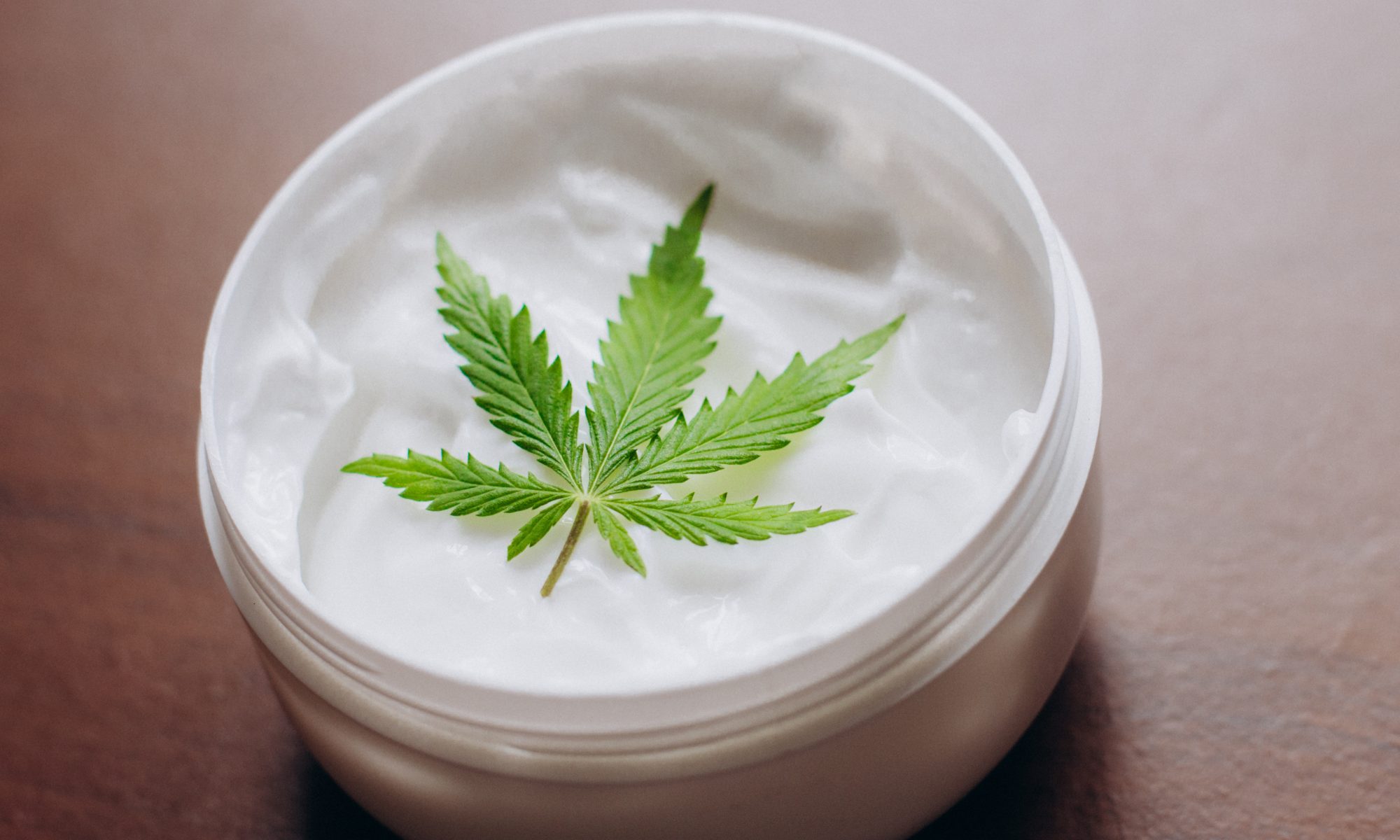 Open jar of white topical cream featuring a cannabis leaf, representing CBD- or cannabis-infused skincare and wellness products.