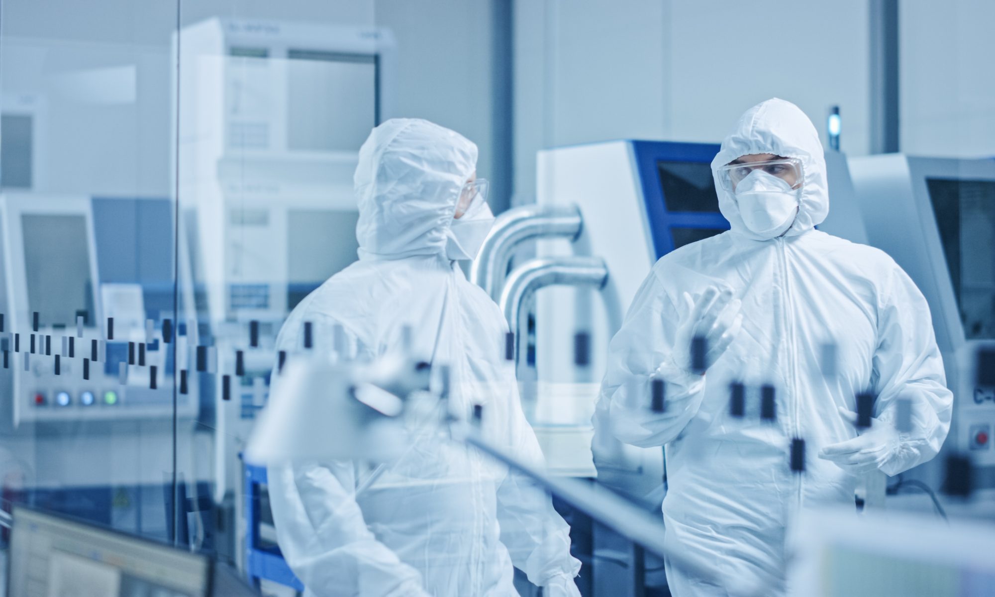 Two technicians in full cleanroom protective suits and masks confer inside a sterile biomanufacturing facility with advanced processing equipment.