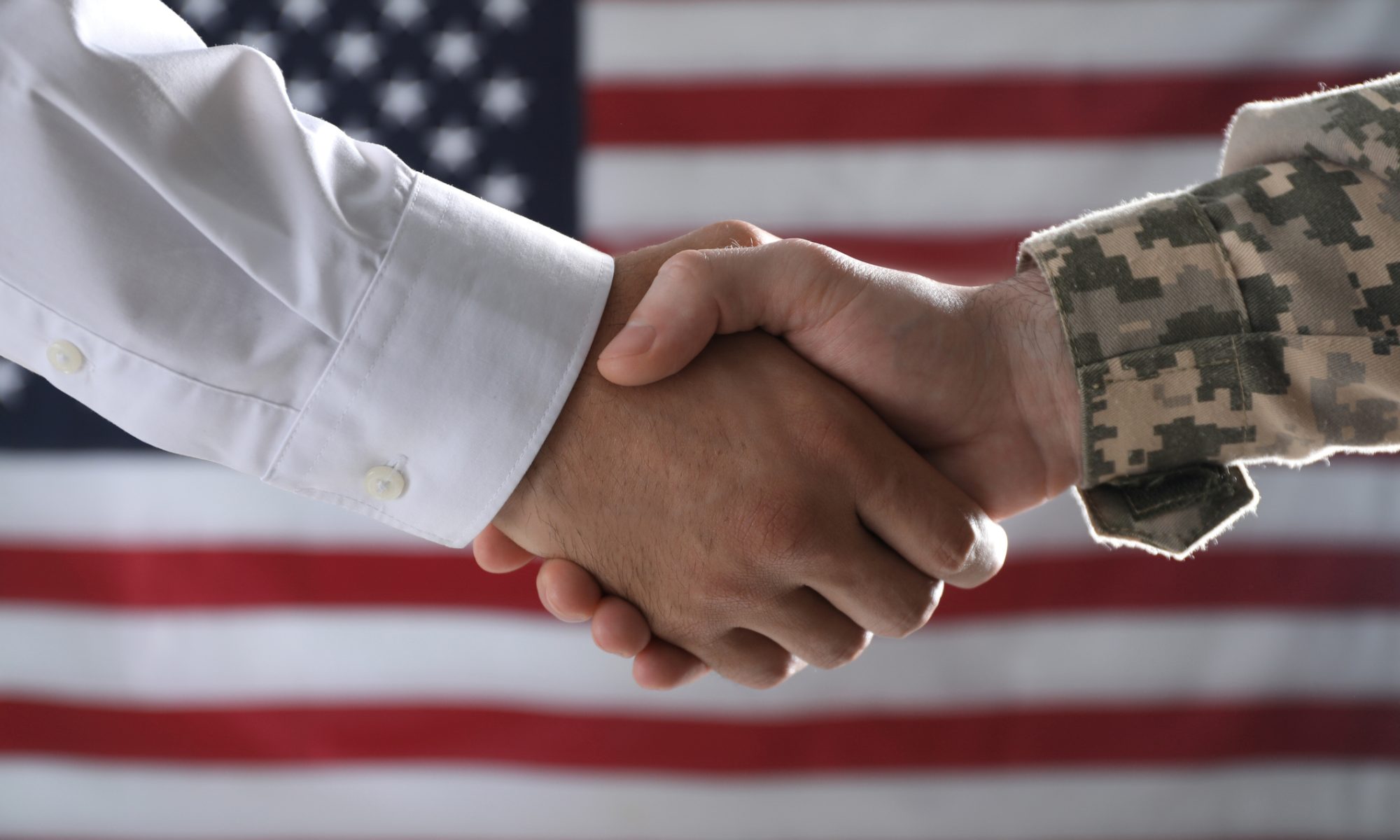 Business professional and U.S. military service member shaking hands in front of an American flag, symbolizing a defense contract partnership.