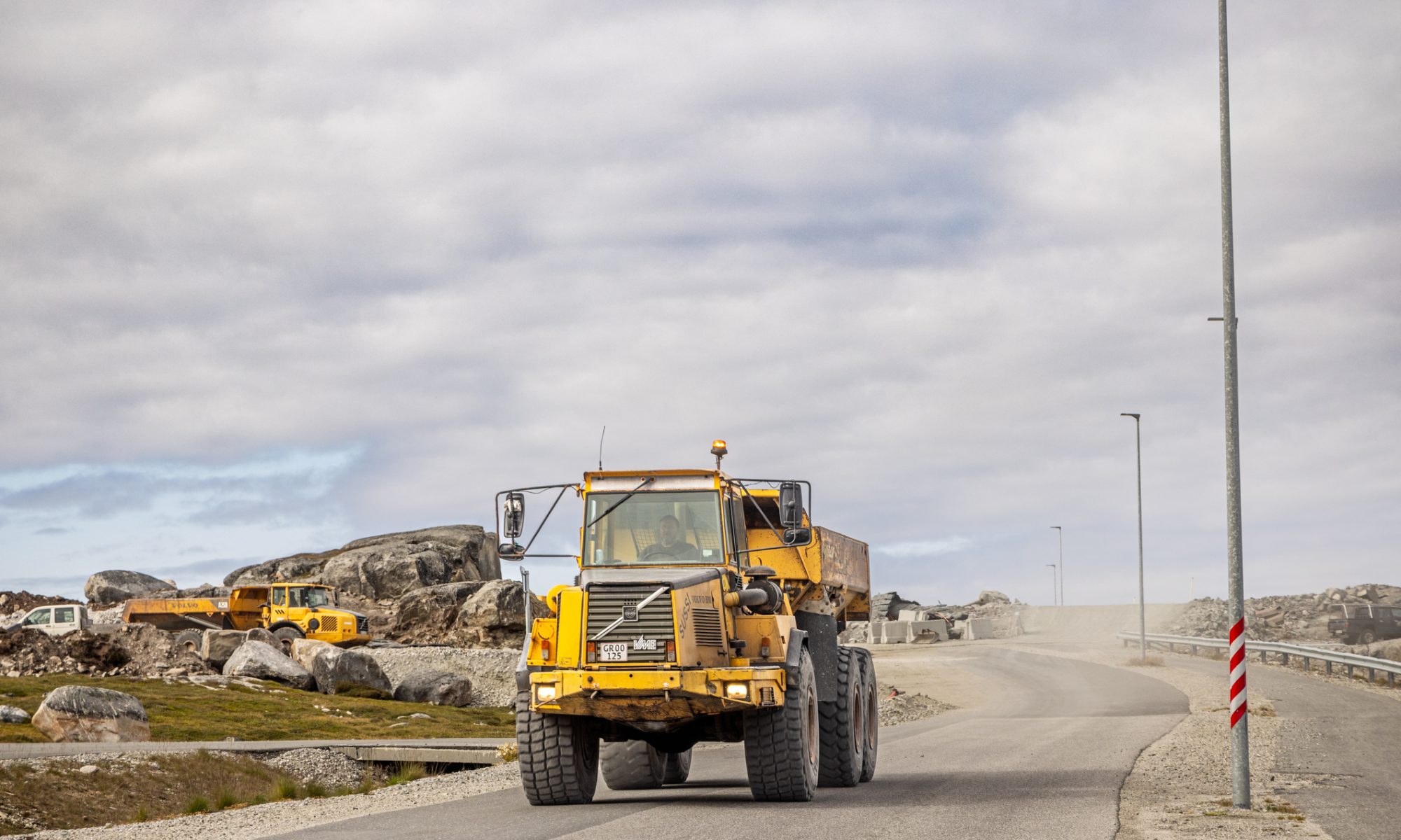 Heavy mining haul truck traveling along a gravel road at an active construction site in Greenland, with rocky terrain and industrial equipment in the background.