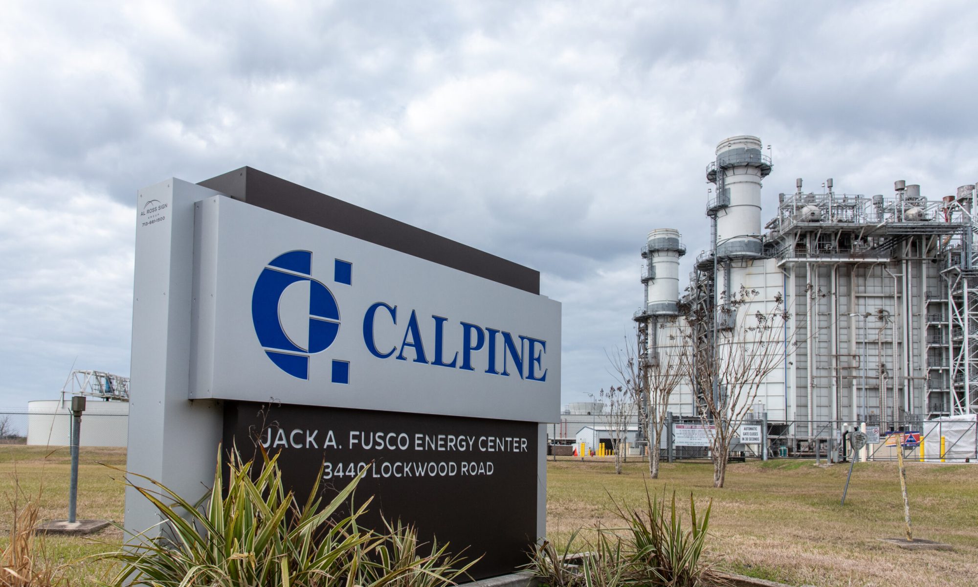 Sign for Calpine’s Jack A. Fusco Energy Center in front of a large natural gas power plant with industrial stacks and processing units under a cloudy sky.