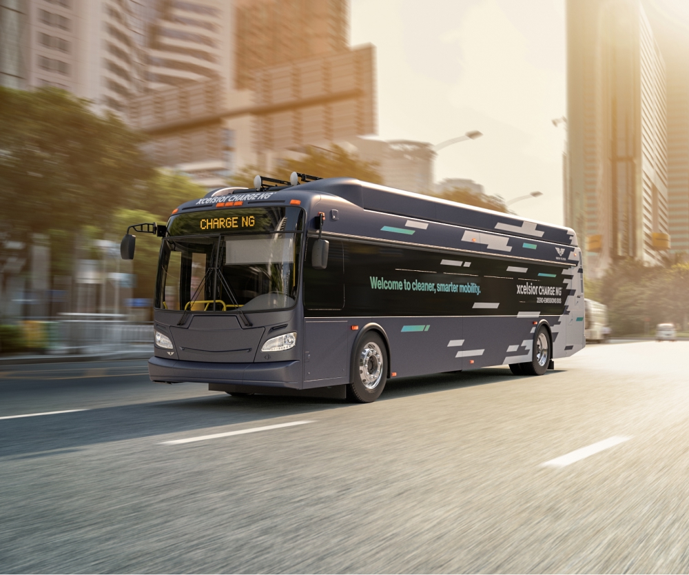 Modern low-emission transit bus driving through a city street, with clean-energy branding on the side and digital destination signage, surrounded by tall buildings and morning sunlight.