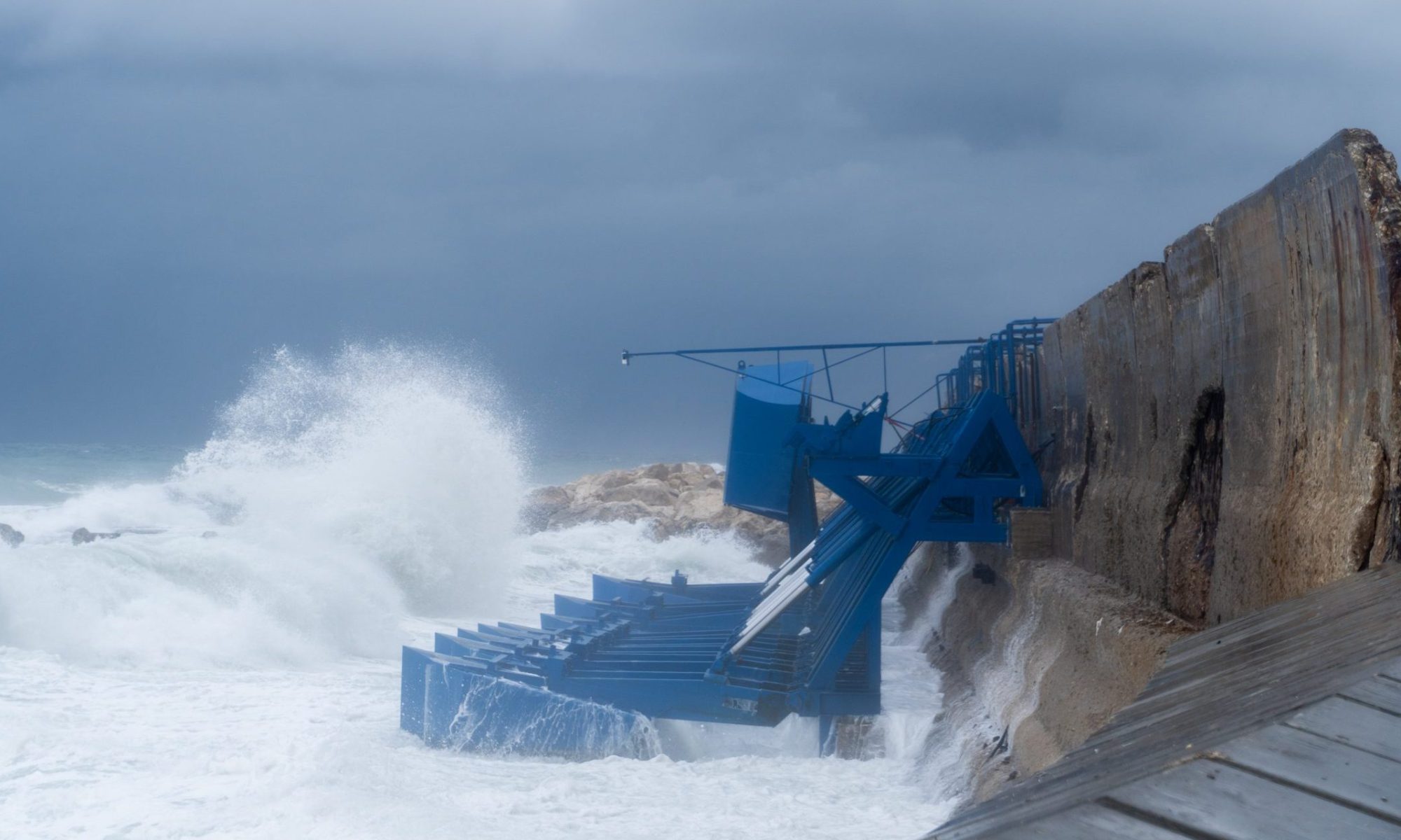 Wave energy conversion equipment mounted along a seawall as ocean waves crash against the structure.
