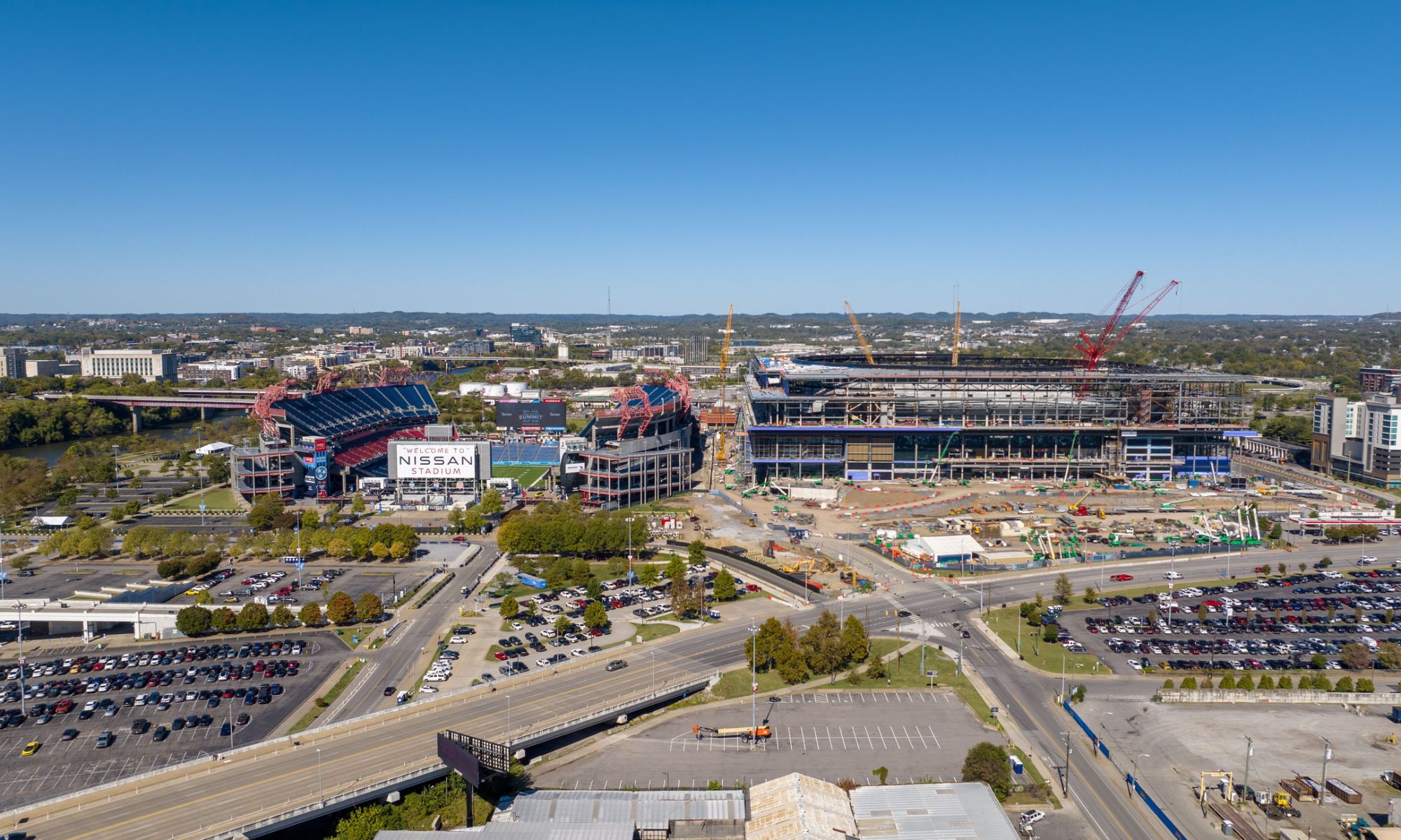 An aerial view of the new Nissan Stadium under construction in Nashville, Tennessee, next to the existing stadium, with cranes, construction equipment, and surrounding city infrastructure visible.