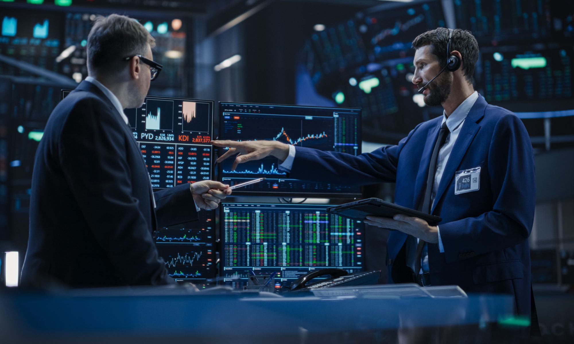 Two financial professionals analyze live stock market data on multiple trading monitors inside a modern trading floor, discussing intraday price movements and market trends.