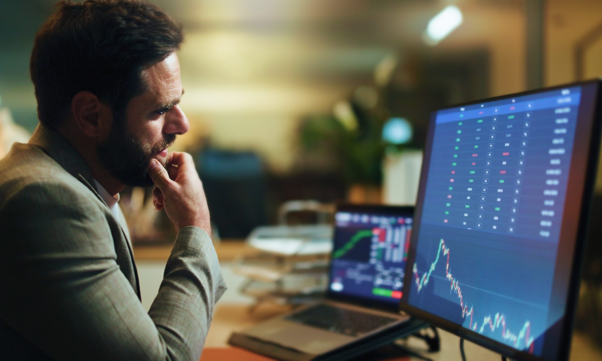 An investor studies stock market charts and a live trading dashboard on multiple computer monitors, representing active market analysis and intraday trading.