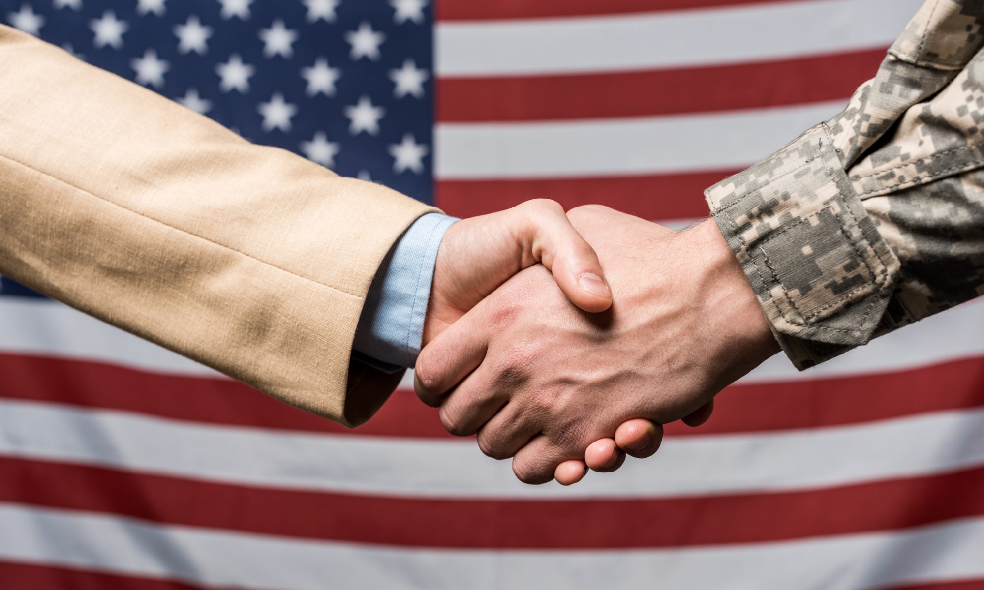 A business executive and a military service member shake hands in front of a U.S. and Canadian flag-themed backdrop, symbolizing a major government defense and satellite communications agreement.