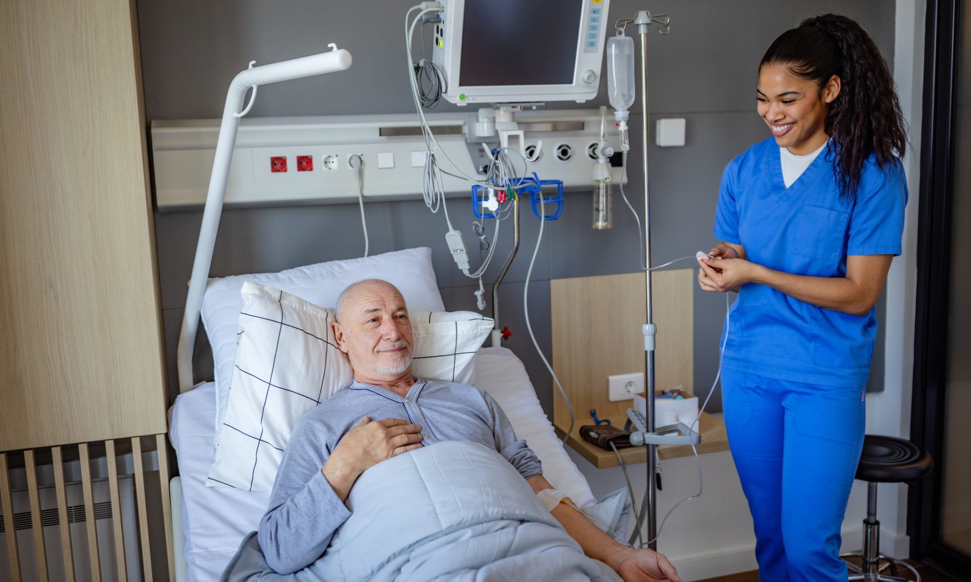 A healthcare professional stands beside a patient receiving treatment in a hospital room, representing the start of a clinical trial for a new prostate cancer immunotherapy.