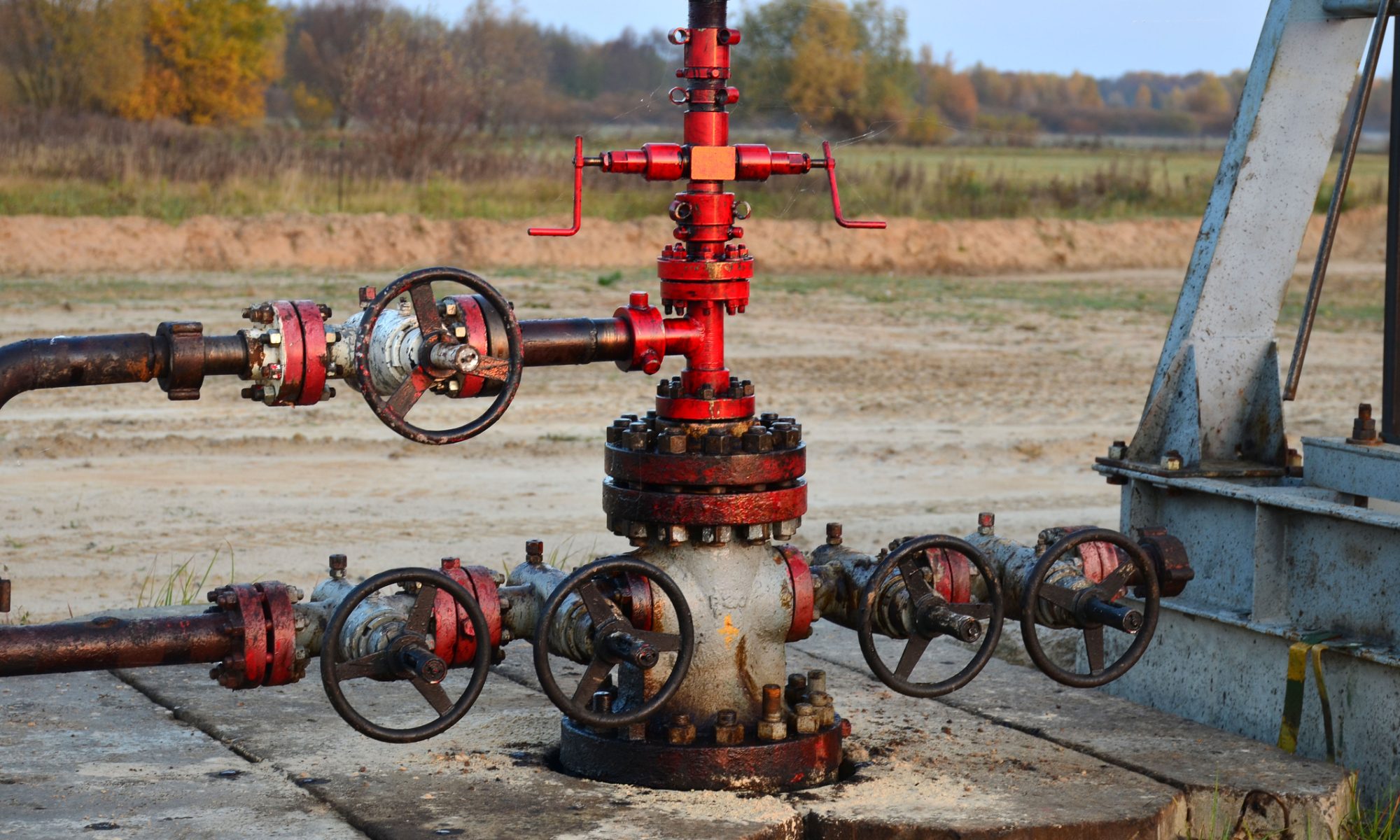 An oil wellhead with red valves and pipeline equipment at an onshore drilling site, representing oil production and upstream energy assets.