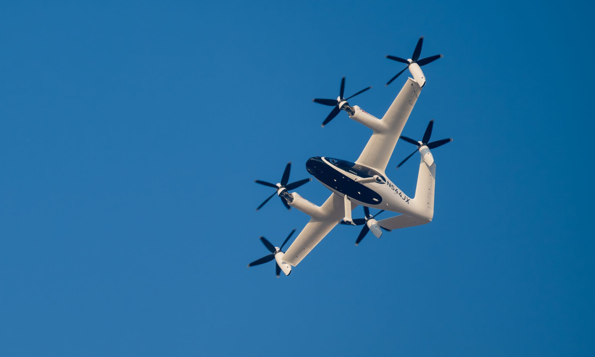 Joby Aviation electric vertical takeoff and landing aircraft flying against a clear blue sky, illustrating the company’s expansion into defense technology through its planned $500 million acquisition of Resonant Sciences.