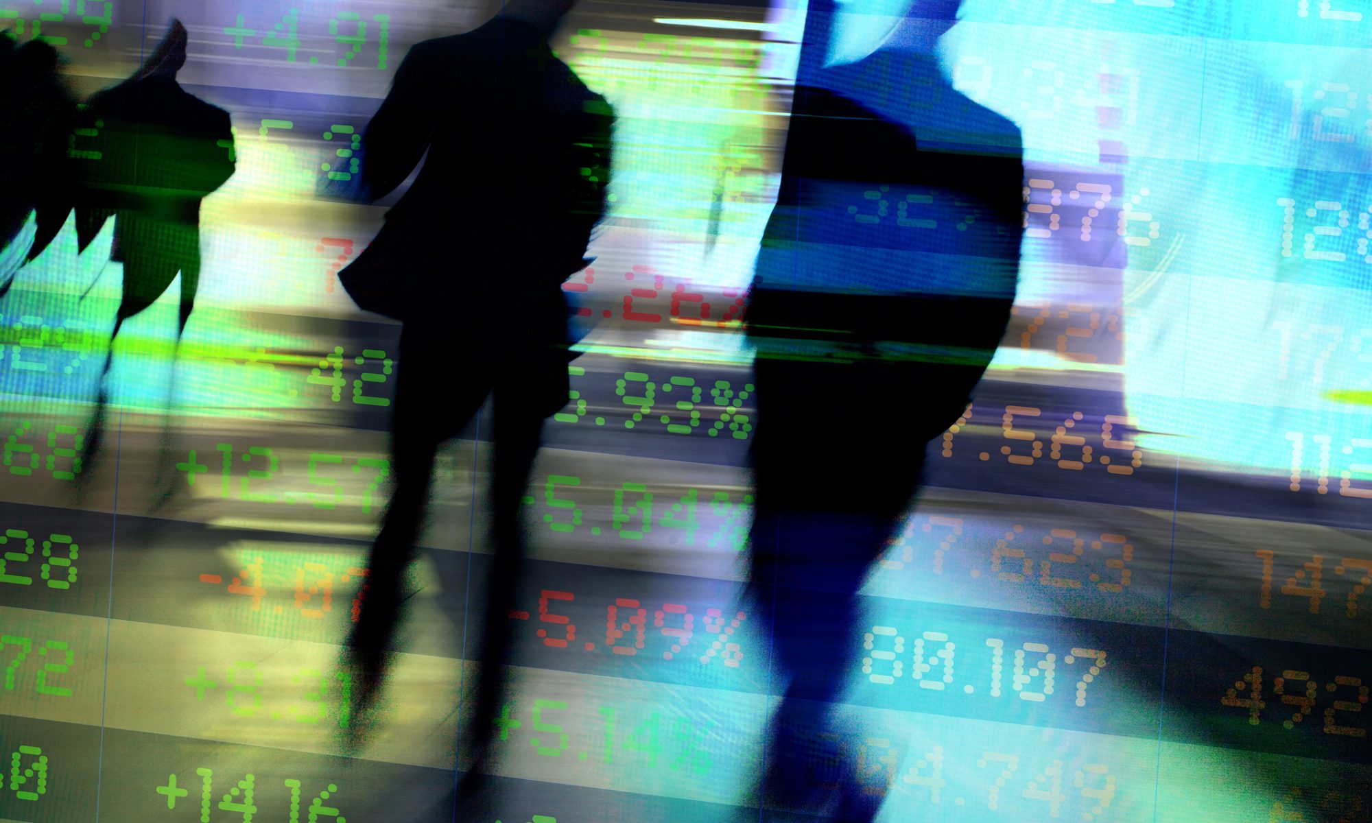 Silhouettes of people walking in front of a digital stock market display showing rising and falling market data, representing active trading in EV and communications and media stocks.