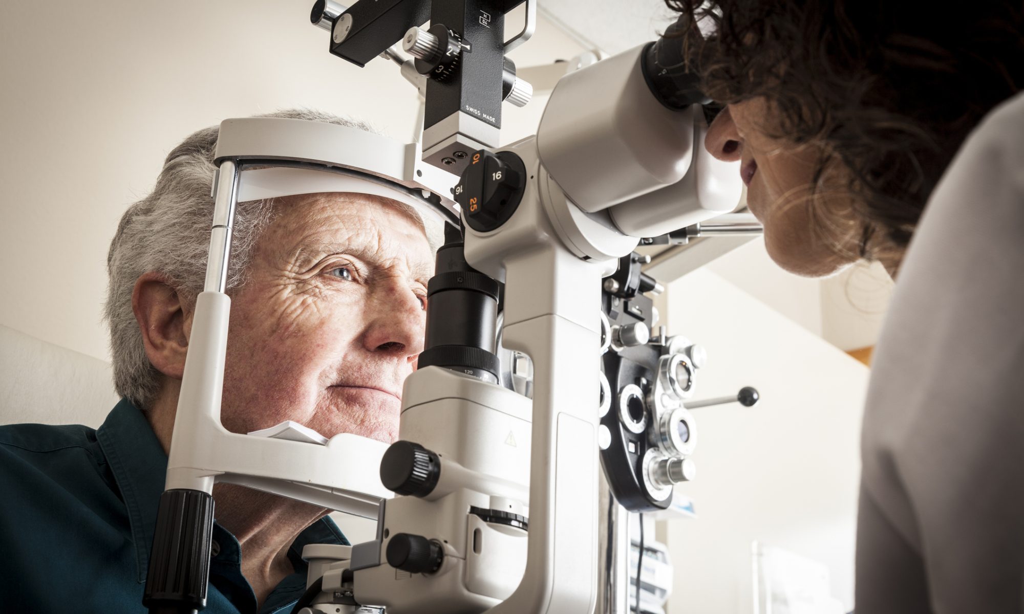 Eye doctor examining an older patient with ophthalmic equipment, representing clinical research into treatments for geographic atrophy and age-related macular degeneration.