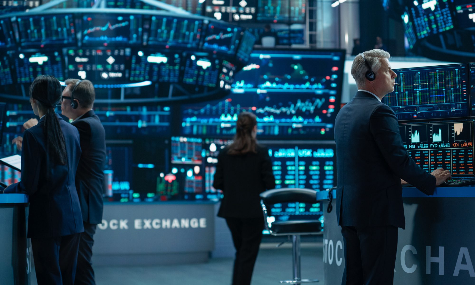 Traders monitor stock charts and market data across multiple screens on a busy exchange floor, representing heavy trading activity in biotech and consumer products stocks.