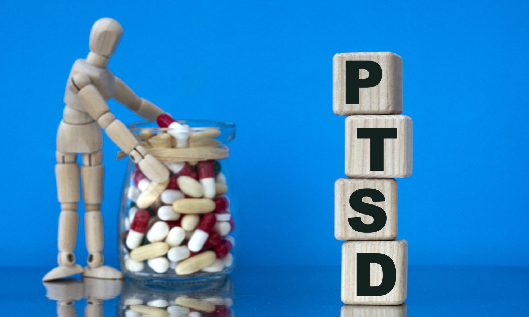 Wooden figure beside a jar of medication and stacked blocks spelling PTSD, representing research into new treatments for post-traumatic stress disorder.