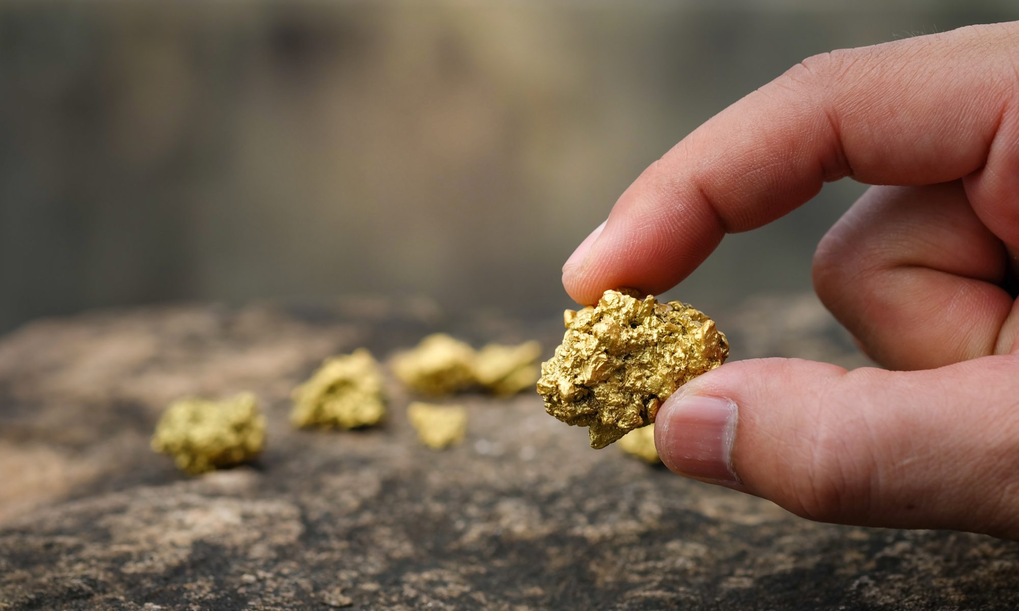 Close-up of a person holding a gold nugget over a rocky surface, representing gold exploration, mineral recovery and mining development.