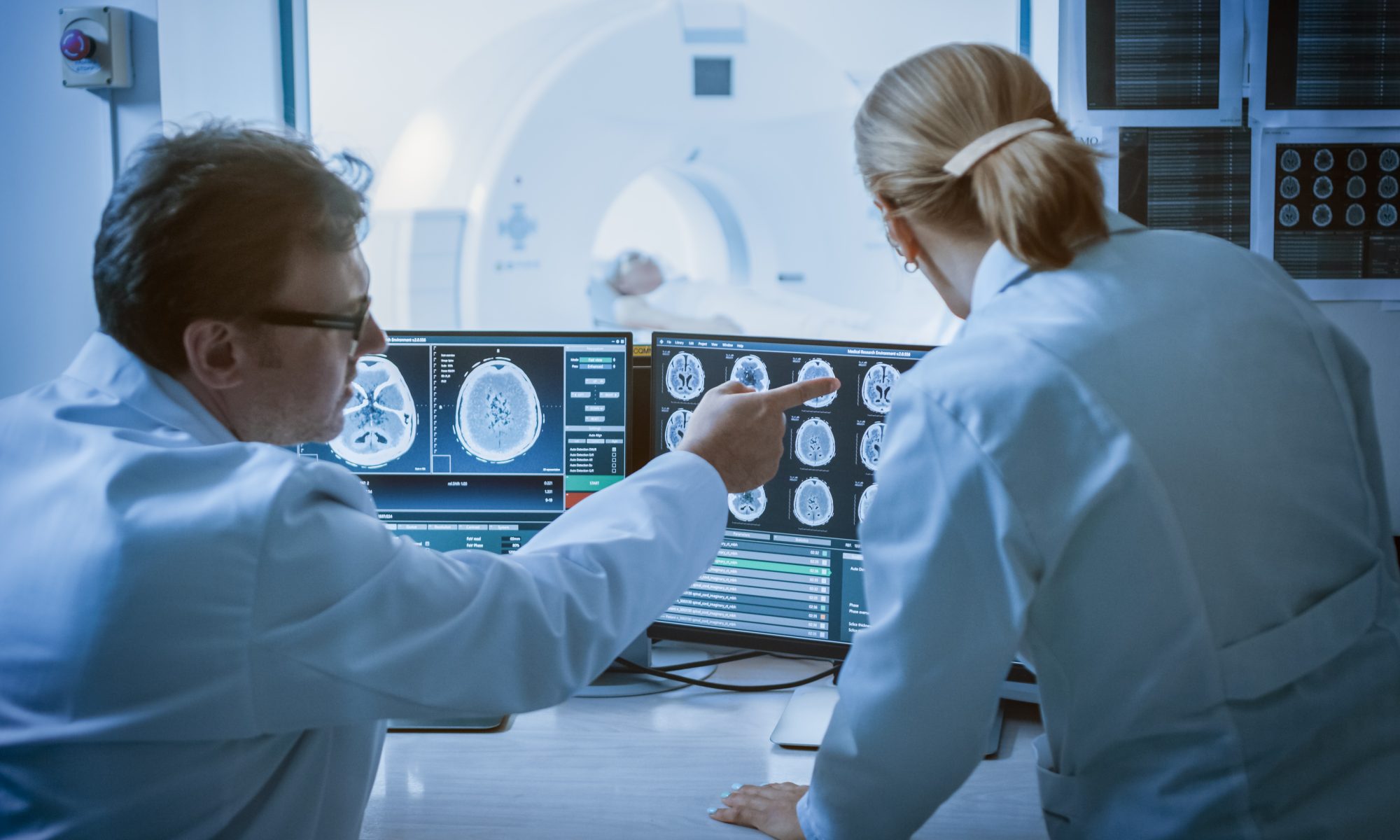 Medical researchers review brain scans on computer monitors as a patient undergoes imaging, representing clinical research into neurological disorders.