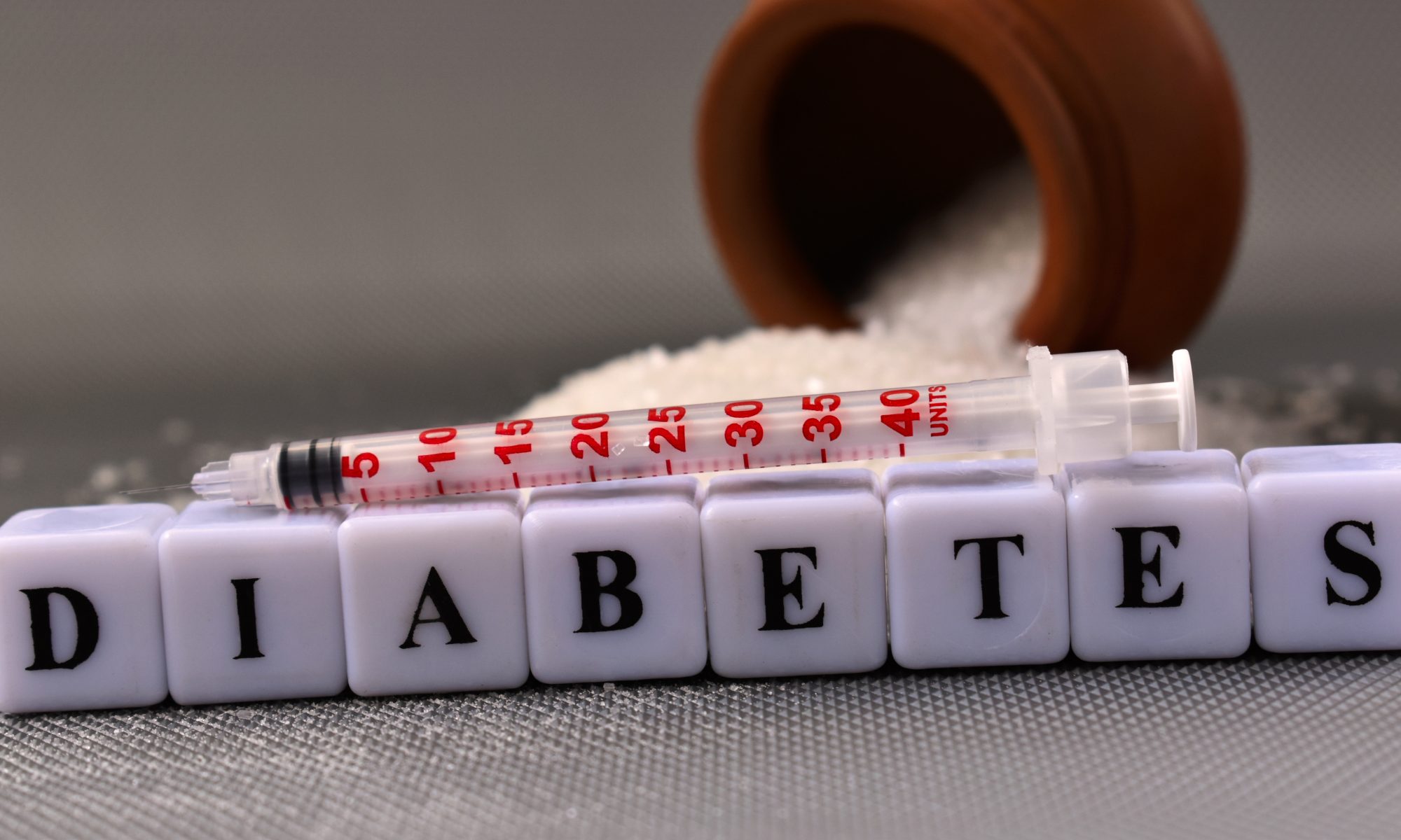 Insulin syringe resting above blocks spelling “diabetes,” with a container of white granules in the background.