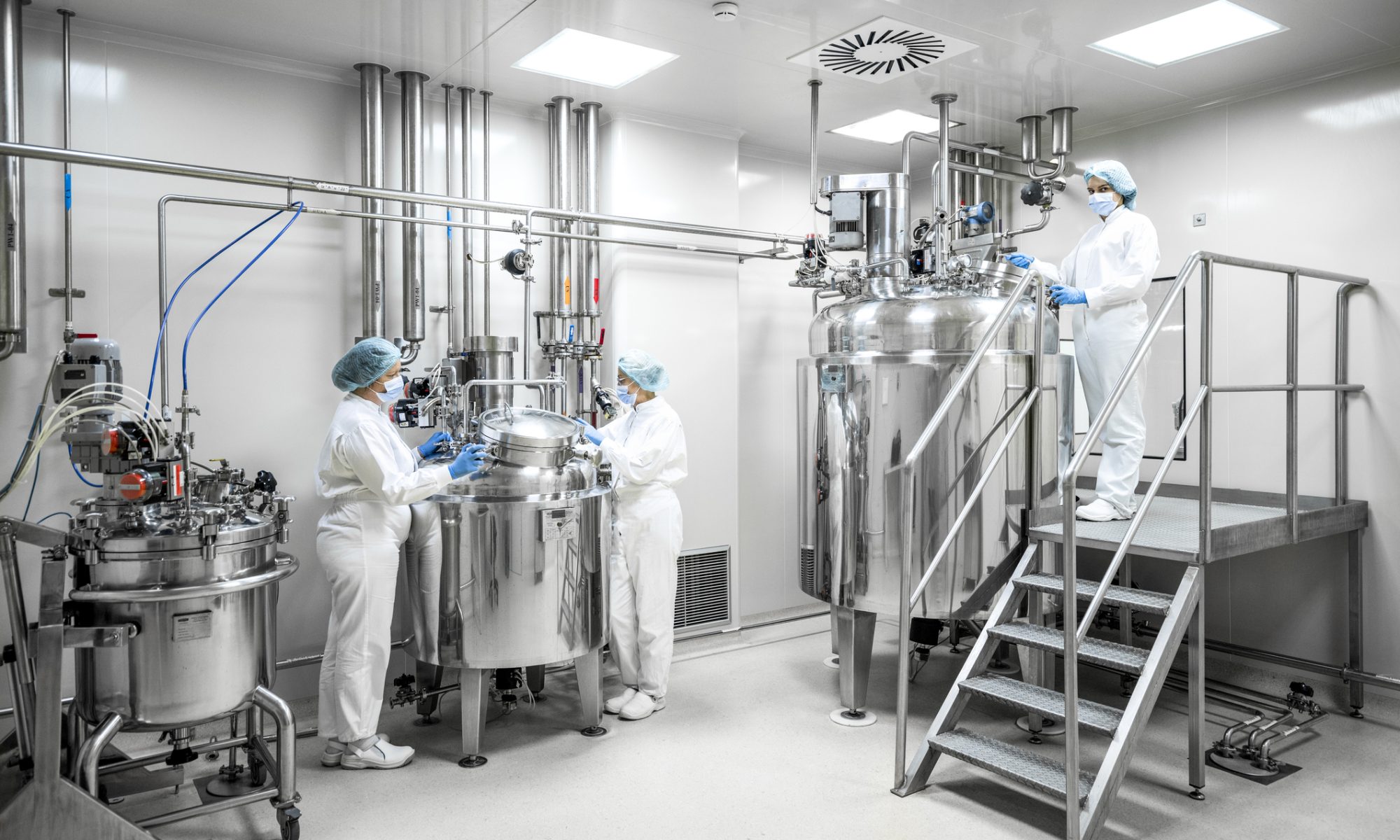 Technicians work with stainless-steel processing equipment inside a pharmaceutical manufacturing facility.