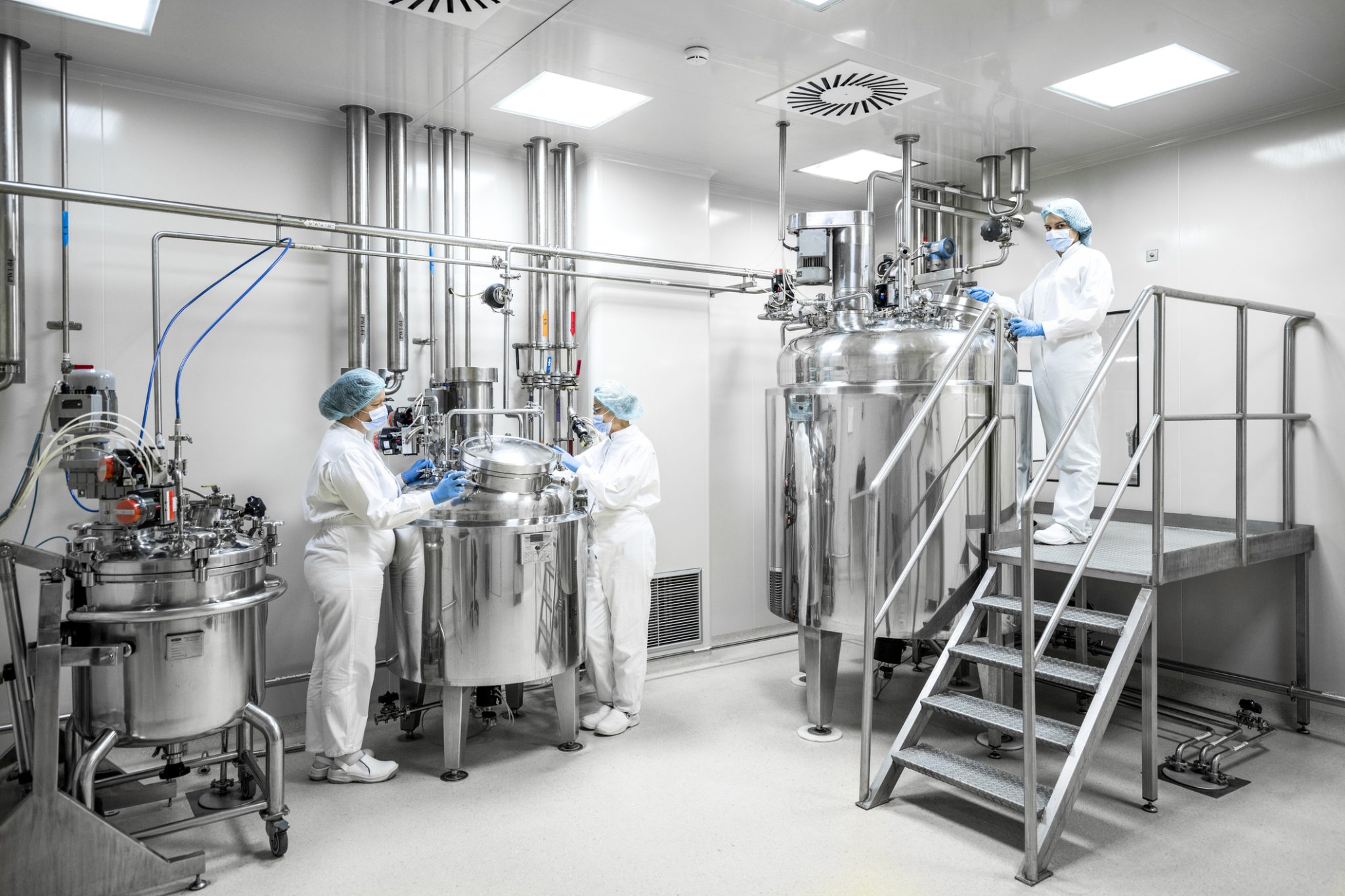 Technicians work with stainless-steel processing equipment inside a pharmaceutical manufacturing facility.
