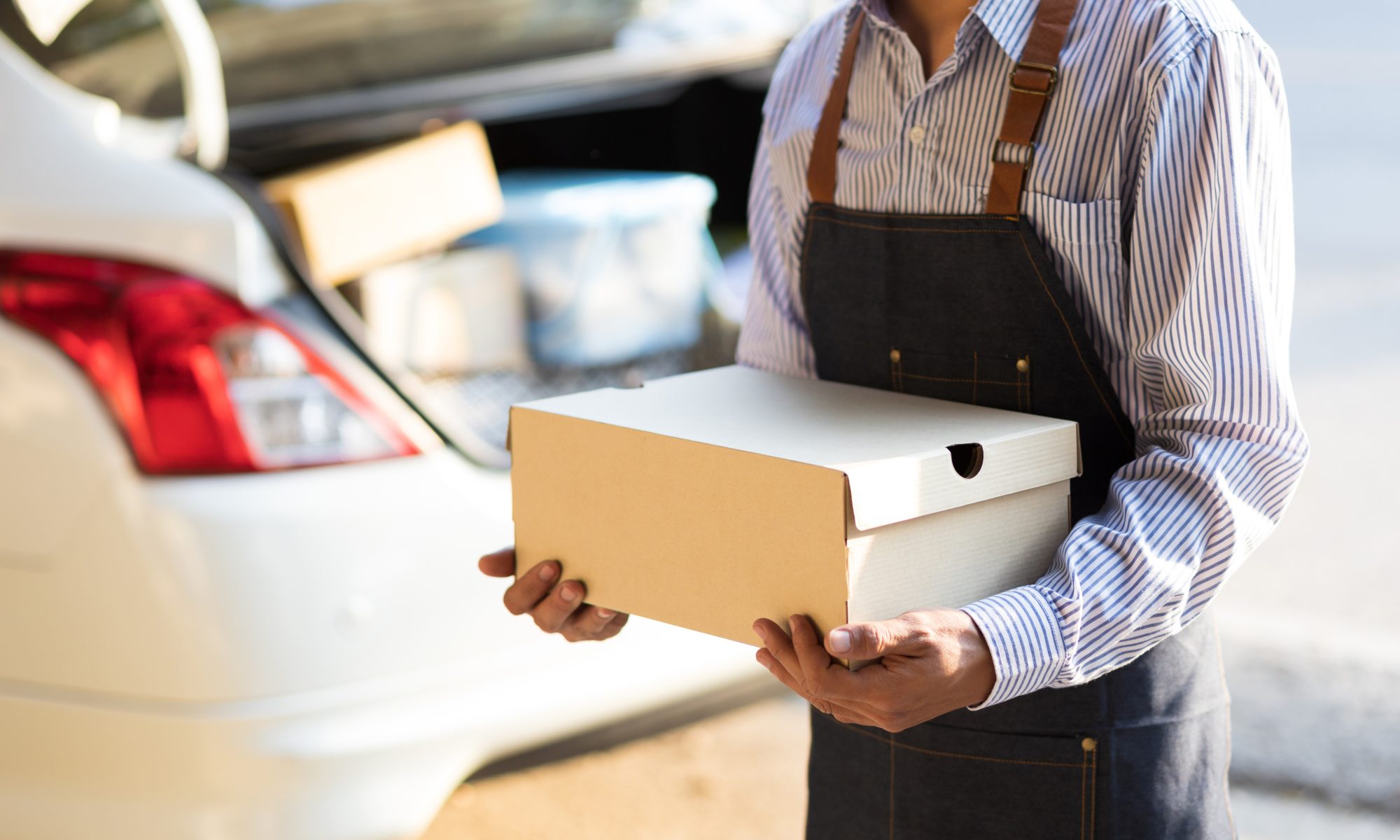 Catering worker carries a food delivery box beside a vehicle, representing workplace catering and large-order food delivery.