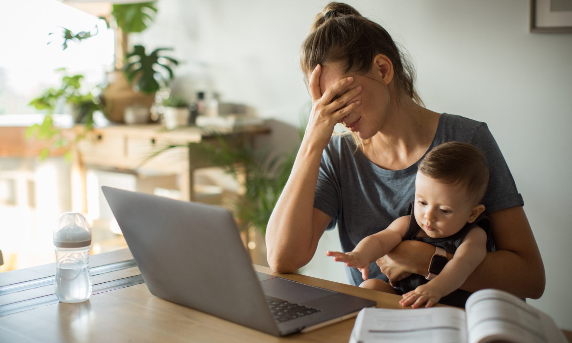 A tired mother holds her baby while sitting at a table with a laptop, illustrating the emotional and mental health challenges parents can face.