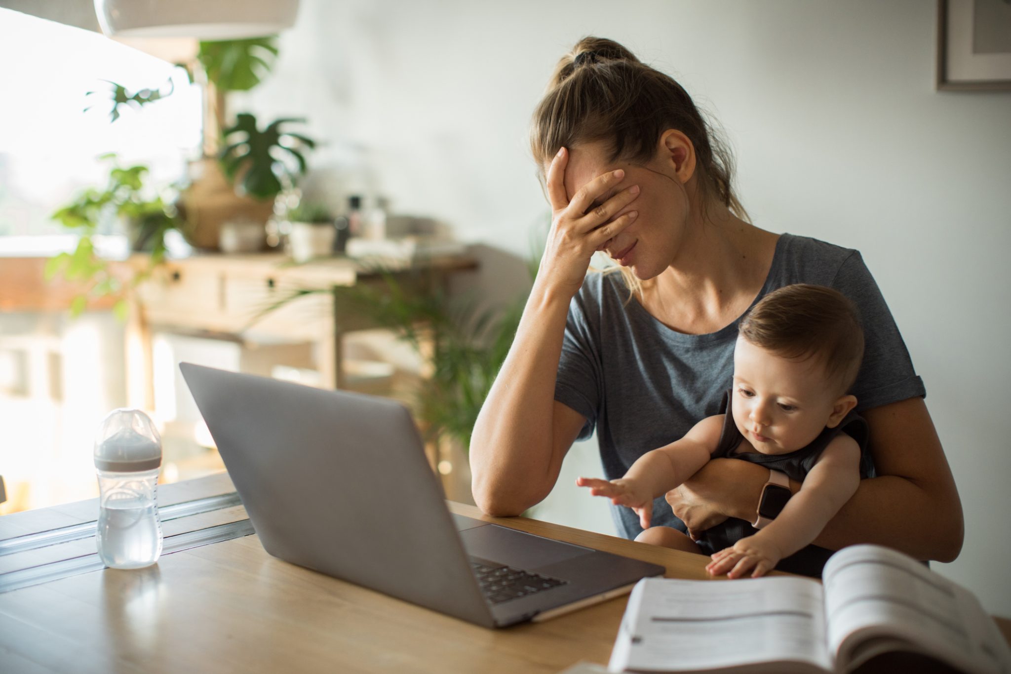 A tired mother holds her baby while sitting at a table with a laptop, illustrating the emotional and mental health challenges parents can face.