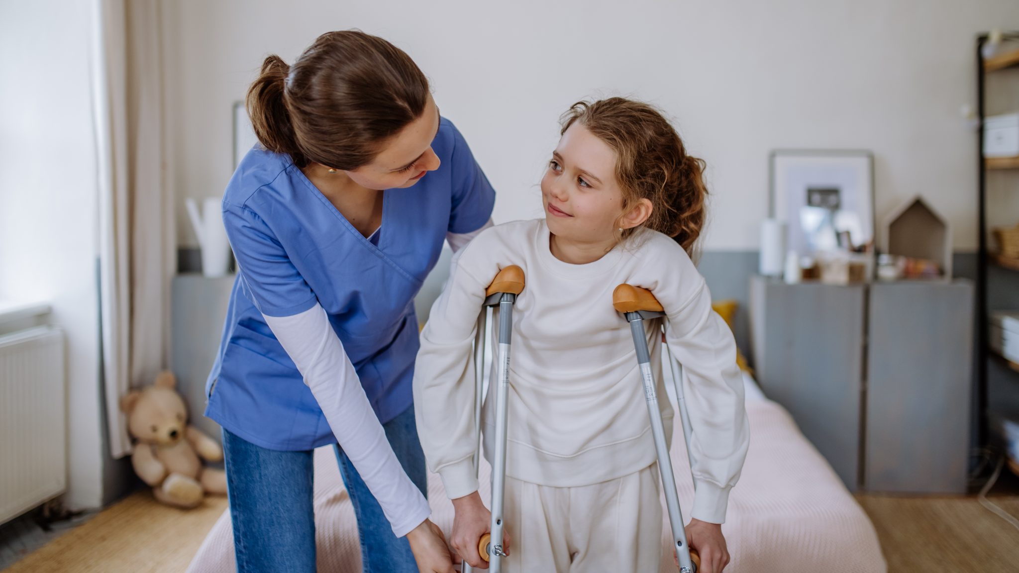 Healthcare professional in blue scrubs assisting a young girl using crutches, illustrating pediatric mobility challenges and treatment needs associated with rare bone diseases.