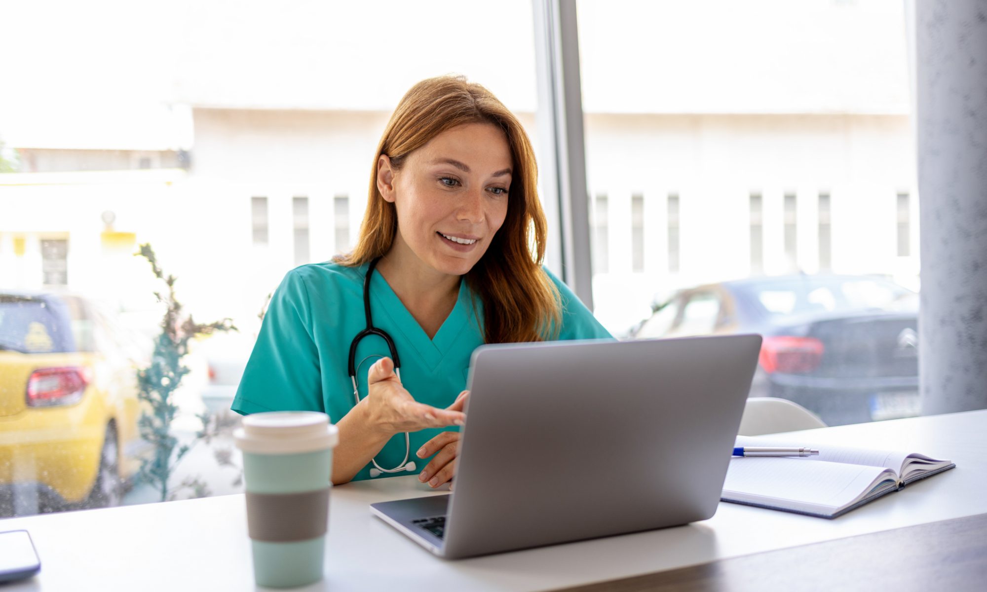 Female healthcare professional wearing teal scrubs and a stethoscope speaks through a laptop, illustrating virtual healthcare and telehealth services for women.