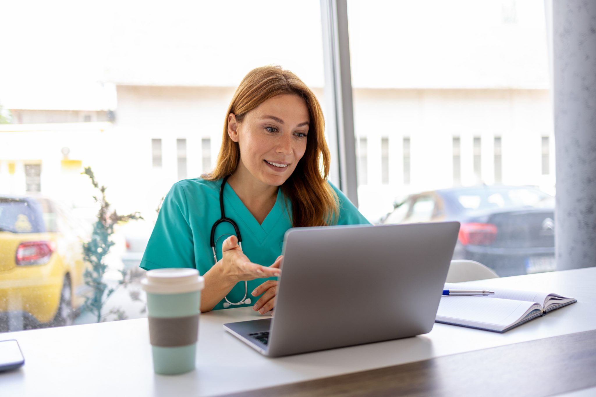 Female healthcare professional wearing teal scrubs and a stethoscope speaks through a laptop, illustrating virtual healthcare and telehealth services for women.