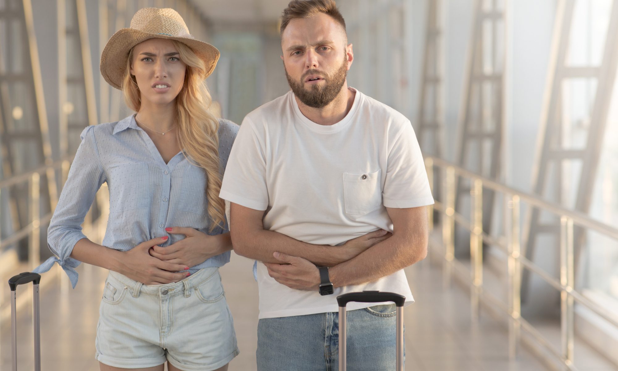 Two travelers holding their stomachs in discomfort while standing with luggage, illustrating travelers’ diarrhea and Immuron’s research into Travelan as a potential preventive treatment.