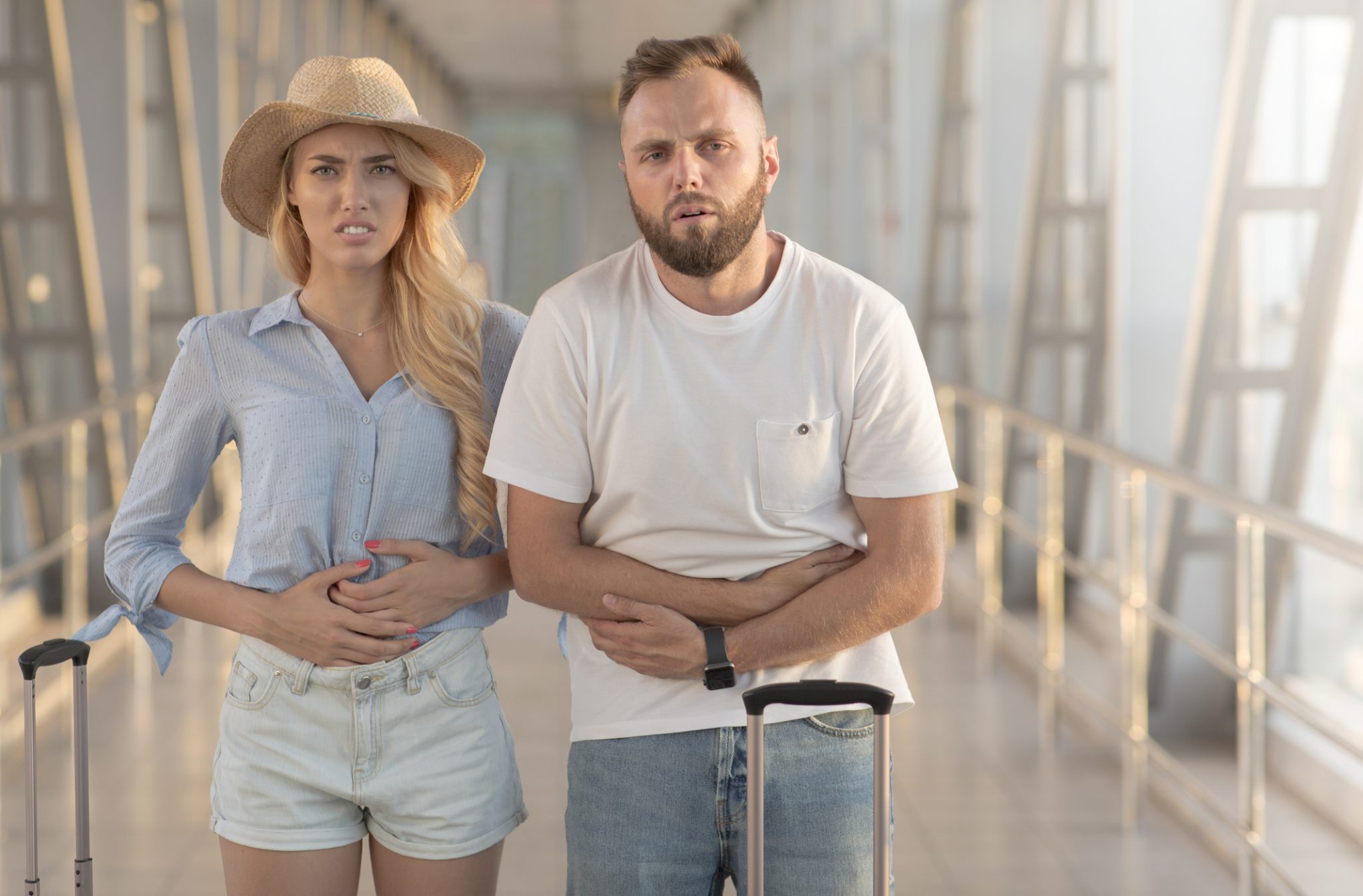 Two travelers holding their stomachs in discomfort while standing with luggage, illustrating travelers’ diarrhea and Immuron’s research into Travelan as a potential preventive treatment.