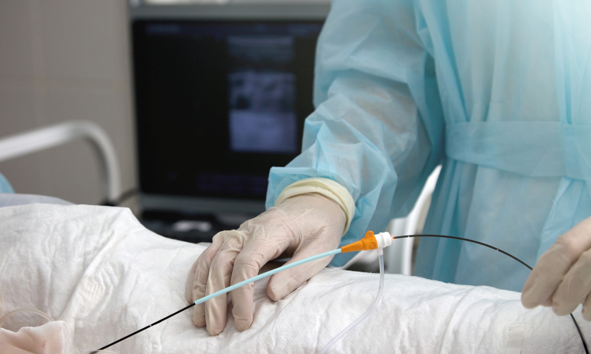 Medical professional wearing surgical gloves and a blue gown performing a minimally invasive procedure using a catheter-like device, with ultrasound equipment in the background.