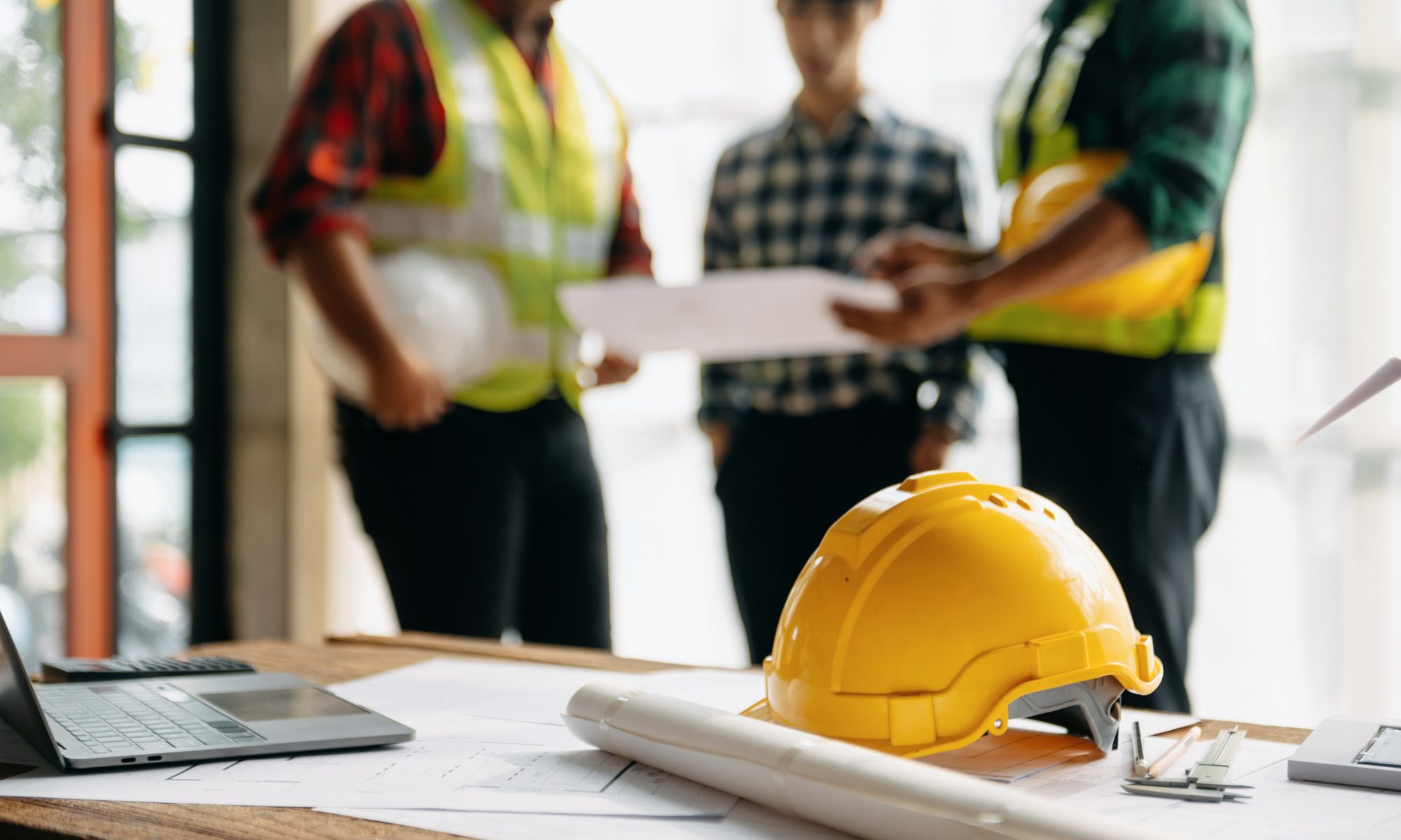 Yellow construction hard hat resting on architectural blueprints and building plans, with construction professionals reviewing documents in the background, representing residential development and smart-home technology integration.