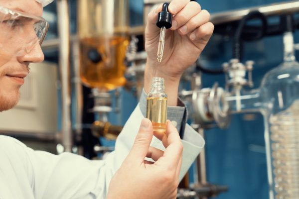Scientist wearing safety glasses and a lab coat uses a dropper to examine a liquid sample in a laboratory, illustrating pharmaceutical research and drug development.