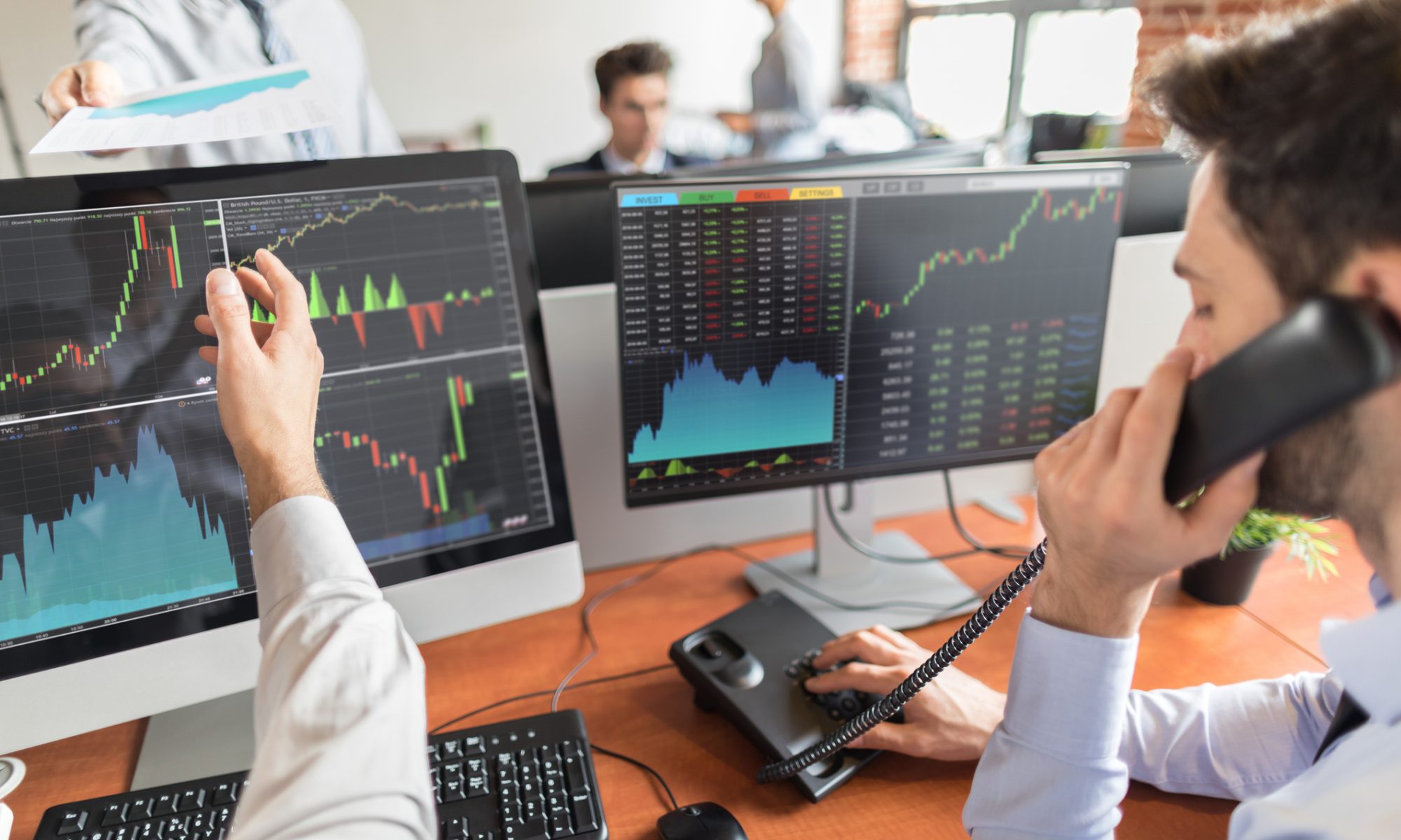 Stock market traders monitor financial charts and market data on multiple computer screens while discussing trades in a busy trading office.
