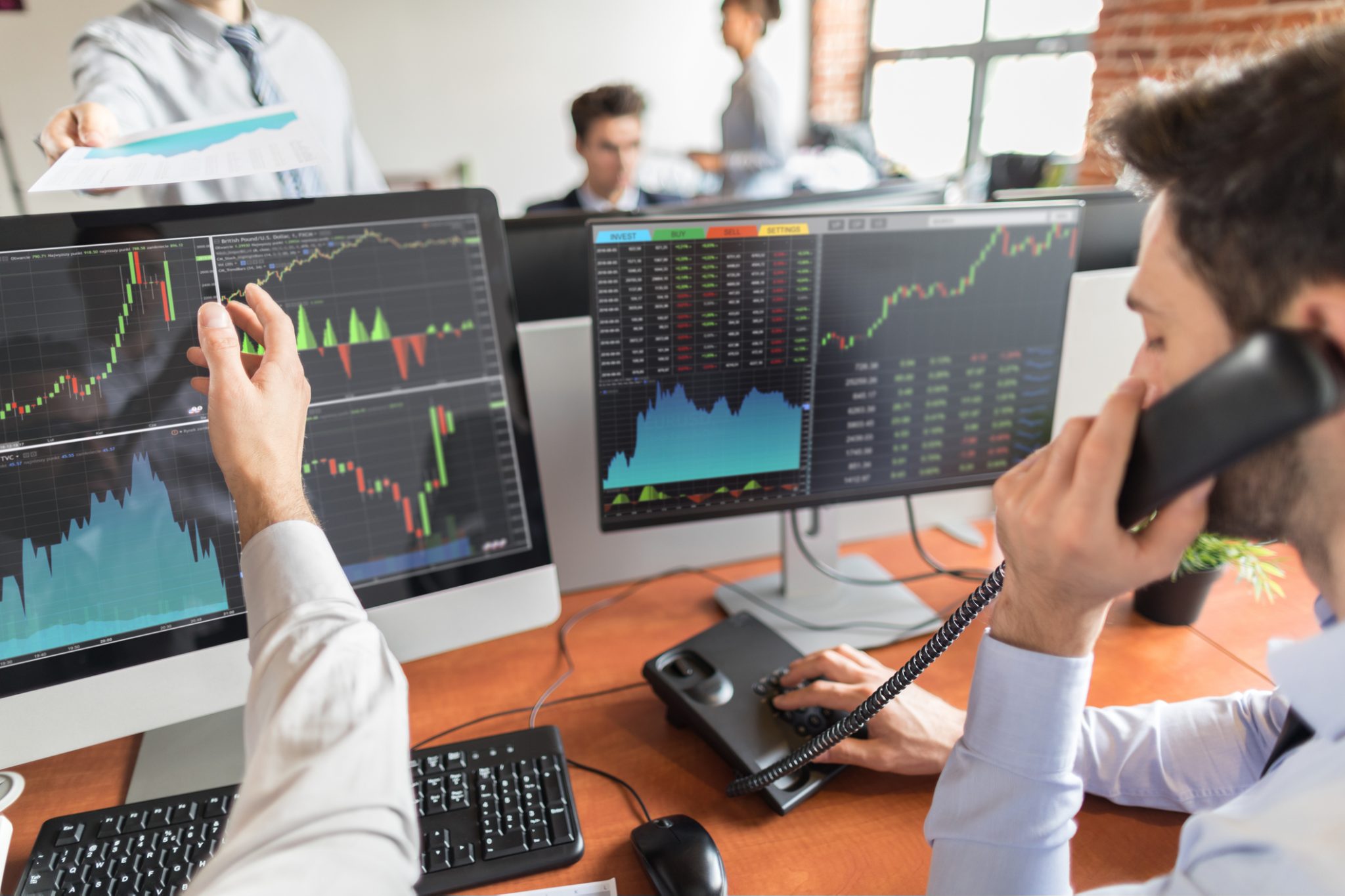 Stock market traders monitor financial charts and market data on multiple computer screens while discussing trades in a busy trading office.